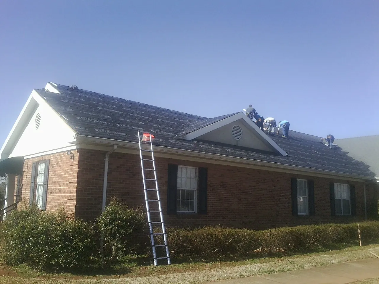 Crew tearing shingles off a brick ranch roof with a ladder set — Emergency Roof Repair work by our crew in Lewisville